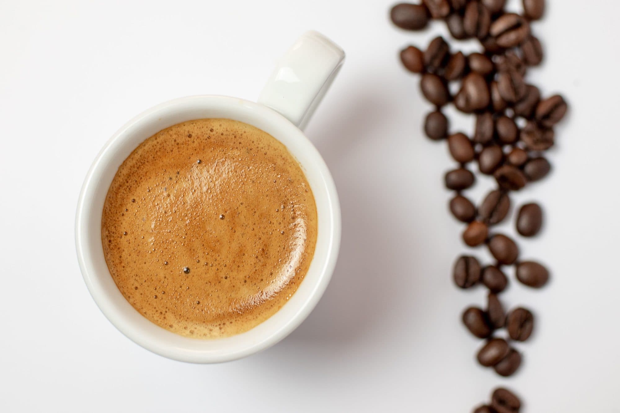 Espresso cu cremă densă într-o ceașcă albă, lângă boabe de cafea prăjite, văzut de sus