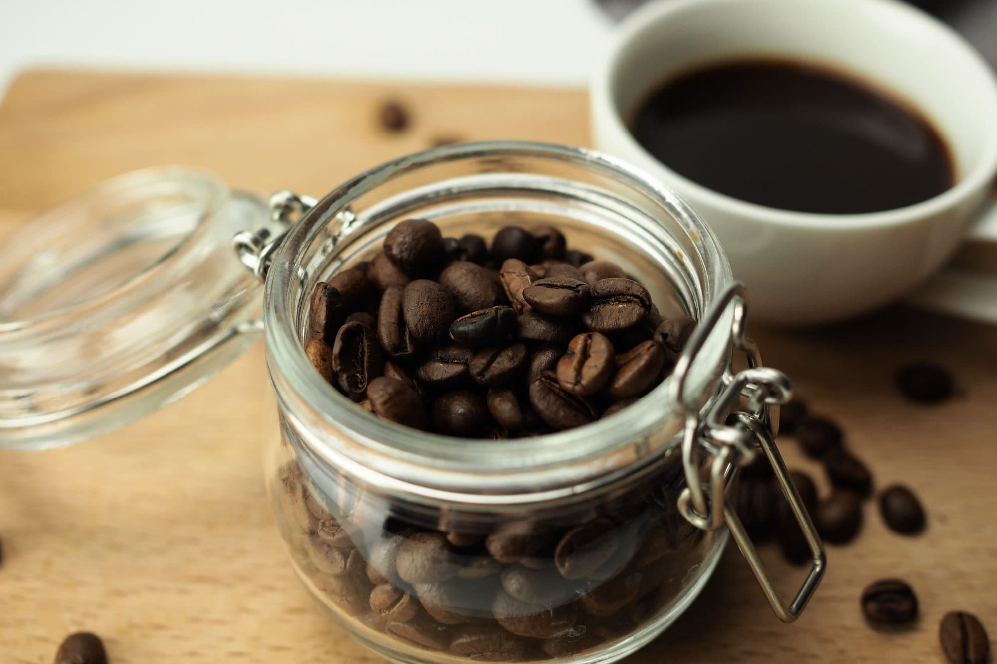 Borcan de sticlă ermetic, cu clemă, plin cu boabe de cafea prăjite, pe un blat de lemn lângă o ceașcă de cafea
